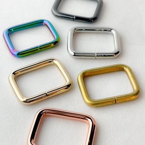May include: Five square-shaped metal rings in different colors: black, iridescent green, silver, gold, and rose gold. The rings are all the same size and have a smooth finish.
