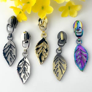 May include: A collection of five zipper pulls with leaf-shaped charms. The pulls have various metallic finishes, including silver, gold, and iridescent. The leaves have intricate vein details, and the pulls are displayed against a white background.