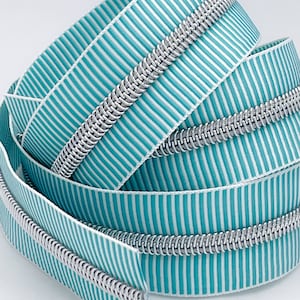 May include: Close-up of a teal and white striped zipper tape with a silver zipper coil. The zipper tape is folded and coiled, showing the detail of the zipper.
