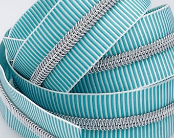 May include: Close-up of a teal and white striped zipper tape with a silver zipper coil. The zipper tape is folded and coiled, showing the detail of the zipper.