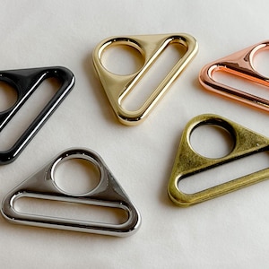 May include: Five triangle-shaped metal rings in different finishes: black, silver, gold, copper, and brass. Each ring has a rounded opening and a flat, rectangular bar.