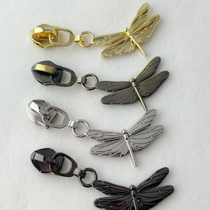 May include: Four metal zipper pulls with dragonfly charms. The zipper pulls are in gold, gunmetal, silver, and black. The dragonfly charms are detailed with wing and body features.