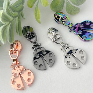 May include: Four different colored metal zipper pulls shaped like ladybugs. The ladybugs are in silver, black, rose gold, and iridescent.