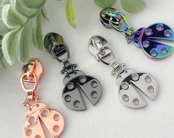 Könnte beinhalten: Vier verschiedene farbige Metall-Reißverschluss-Zipper in Form von Marienkäfern. Die Marienkäfer sind in Silber, Schwarz, Roségold und Regenbogenfarben.