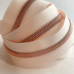 May include: Close-up of a rose gold zipper tape with a white woven fabric backing. The tape is coiled and shows the detail of the zipper teeth.