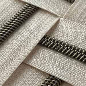 May include: Close-up of a white zipper tape with a brass zipper chain. The zipper tape has a textured surface and the zipper chain is made of metal.