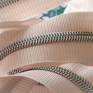 May include: Close-up of a pink zipper tape with a silver metal zipper chain. The tape has a ribbed texture.