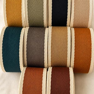 May include: A collection of ten spools of grosgrain ribbon arranged in three rows. Each spool features a solid colored center in shades of mustard, olive, black, beige, teal, gray, tan, orange, rust, and brown, framed by cream-colored ribbed edges.