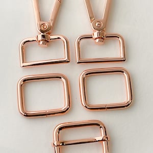 May include: Set of five rose gold metal hardware pieces for crafting. Includes two swivel clasps, two rectangular D-rings, and one rectangular buckle.