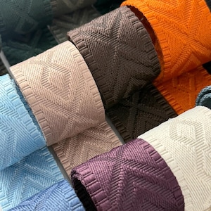 May include: Close-up of several rolls of decorative ribbon in various colors, including teal, orange, brown, and purple. Each ribbon features a textured, geometric pattern, suitable for crafts and embellishments.