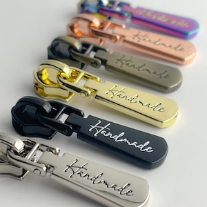 May include: Close-up of six zipper pulls in various metallic finishes: iridescent, copper, bronze, gold, black, and silver. Each pull has the word "Handmade" engraved on it. The pulls are arranged in a row against a white background.