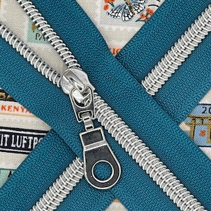 May include: Two teal zipper tapes with silver metal teeth and a silver metal zipper pull. The zipper tapes are crossed over each other.