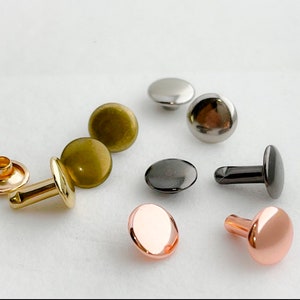 May include: Assortment of metal rivets in various colors, including gold, silver, copper, and black. The rivets are round and have a flat head. They are commonly used for attaching fabric, leather, or other materials.