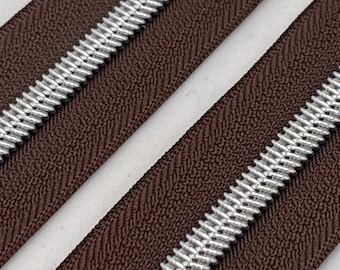 Könnte beinhalten: Nahaufnahme eines braunen Reißverschlussbandes mit silbernen Metall-Reißverschlusszähnen. Das Reißverschlussband hat eine strukturierte Oberfläche.