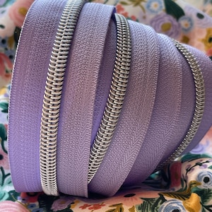 May include: Close-up of a coil zipper in a light purple color. The zipper is made of metal and has a smooth finish.