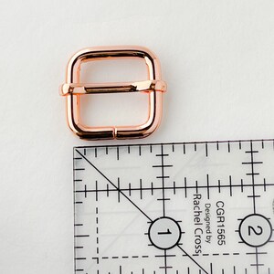 Op de afbeelding: Een roosgoudkleurige metalen gesp voor een band of riem. De gesp is 2,5 cm breed en heeft een vierkante vorm.