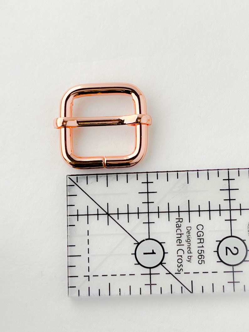 Op de afbeelding: Een roosgoudkleurige metalen gesp voor een band of riem. De gesp is 2,5 cm breed en heeft een vierkante vorm.