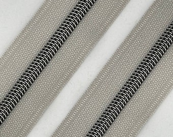 May include: Close-up of a silver zipper tape with a black coil zipper. The zipper tape has a textured surface.