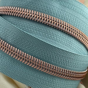Op de afbeelding: Close-up van een turquoise rits met een roségouden spiraal. De ritssluiting heeft een textuur.