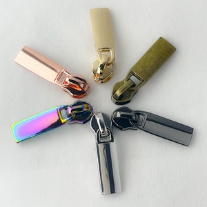 May include: Five zipper pulls in different colors and finishes. The pulls are made of metal and have a rectangular shape. The colors include gold, rose gold, antique brass, iridescent, and black.