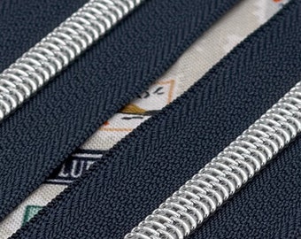May include: Two black zipper tapes with silver metal teeth. The zipper tapes are lying on a fabric background with a pattern of colorful stamps.