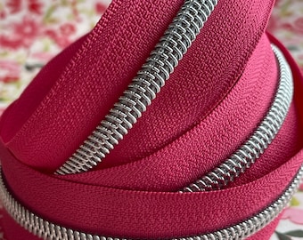 May include: Close-up of a roll of fuchsia-colored zipper tape with a silver metal zipper. The zipper is coiled, revealing the teeth. The background is a floral pattern.