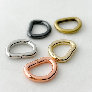 May include: Five D-shaped metal rings in different finishes: black, silver, gold, and copper. The rings are all the same size and are arranged in a semi-circle on a white background.