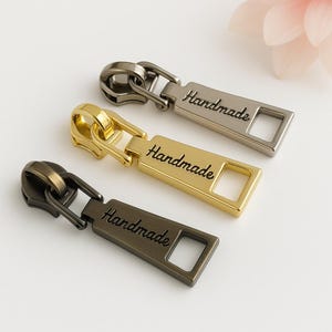 May include: Three metal zipper pulls in bronze, gold, and silver. Each pull has the word "Handmade" engraved on a rectangular plate with a square hole. The zipper pulls are for sewing projects.