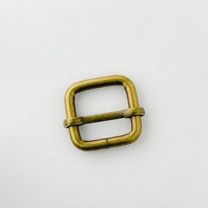 Op de afbeelding: Een messingkleurige metalen gesp met een vierkante vorm. De gesp heeft een gladde afwerking en is ontworpen om te worden gebruikt met een band of riem.