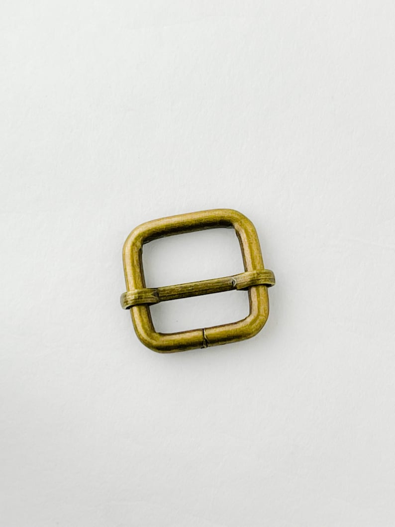 Op de afbeelding: Een messingkleurige metalen gesp met een vierkante vorm. De gesp heeft een gladde afwerking en is ontworpen om te worden gebruikt met een band of riem.