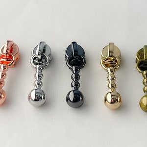 May include: Five zipper pulls with a ball-shaped end. The pulls are in different colors: rose gold, silver, black, gold, and antique brass.