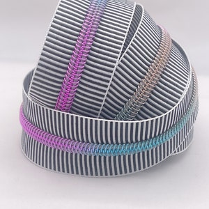 May include: A close-up of three rolls of black and white striped ribbon with iridescent zipper trim. The zipper trim is pink, blue, and green.