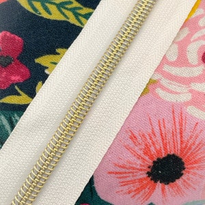 May include: Close-up of a white zipper tape with a gold metal zipper pull. The zipper tape is sewn to a fabric with a floral pattern in shades of pink, red, green, and yellow.