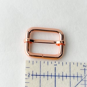 May include: A rose gold metal buckle for a strap or belt. The buckle is about 1 inch wide and is shown next to a ruler.