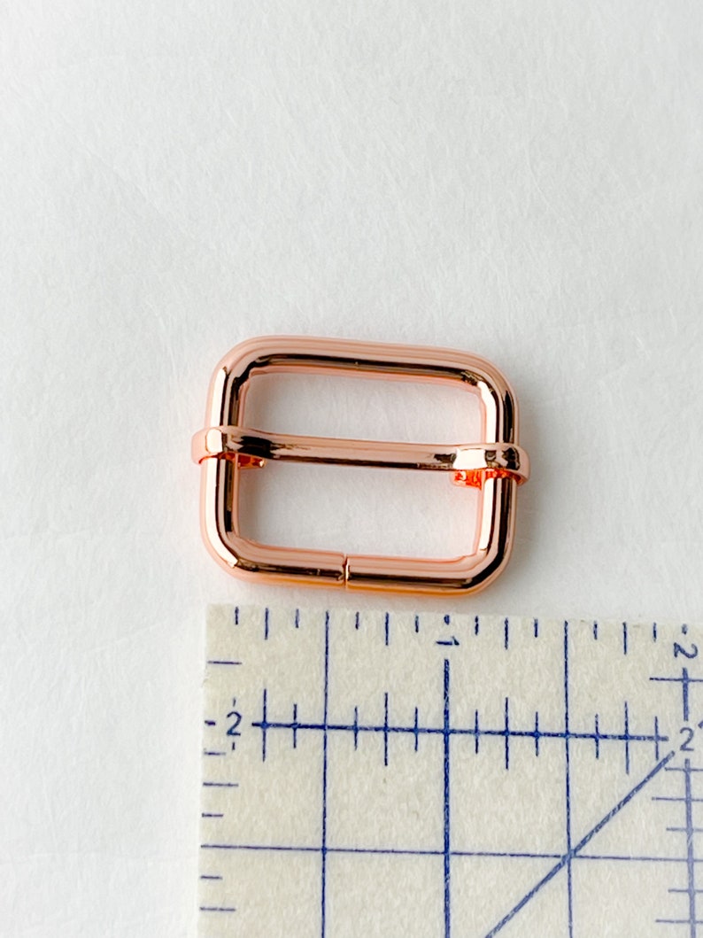 May include: A rose gold metal buckle for a strap or belt. The buckle is about 1 inch wide and is shown next to a ruler.