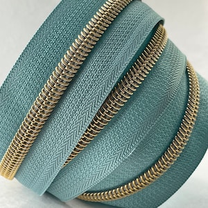 May include: Close-up of a teal zipper with a gold metal zipper pull. The zipper is coiled and folded, showing the texture of the fabric.