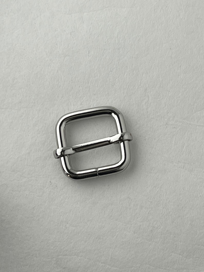 Op de afbeelding: Een zilveren metalen gesp met een vierkante vorm. De gesp heeft een gladde, gepolijste afwerking.