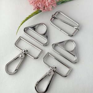 May include: Five silver metal hardware pieces for bags, purses, or other crafts. The pieces include two triangular rings, two rectangular rings, and a swivel clasp.