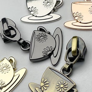 May include: Close-up of several metal zipper pulls. Each pull features a teacup and saucer design with daisy accents. The pulls are in various metallic finishes, including silver, gold, and bronze. The teacups are on saucers.