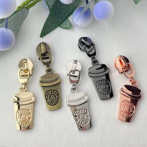 May include: Five zipper pulls with a coffee cup design. The zipper pulls are in gold, silver, black, and rose gold. Each zipper pull has the word "Coffee" written on it.