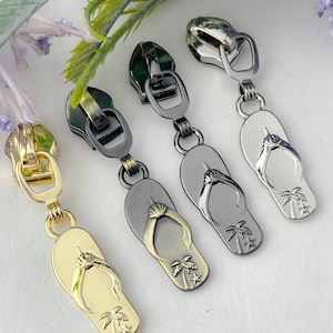 May include: Four zipper pulls in the shape of flip flops with palm trees. The zipper pulls are in gold, silver, black, and antique gold.