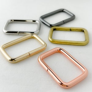 May include: Five metal D-rings in different finishes: silver, black, gold, rose gold, and brass. The rings are all the same size and shape, and they are arranged in a semi-circle on a white surface.
