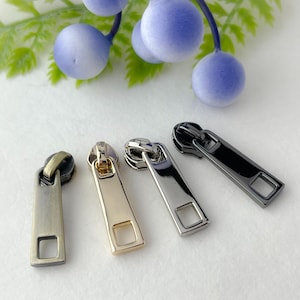 May include: Four zipper pulls in different finishes: antique brass, gold, silver, and black. Each pull has a rectangular shape with a square opening at the bottom.