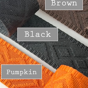 K&ouml;nnte beinhalten: Drei Streifen aus gewebtem Stoffbesatz mit einem geometrischen Rautenmuster. Die Farben sind als Brown, Black und Pumpkin beschriftet. Jeder Streifen weist ein strukturiertes, erhabenes Gewebe mit einem durchgehenden Muster auf.
