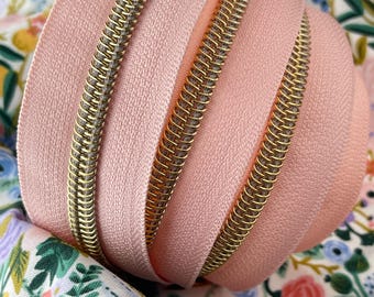 May include: A close-up of a pink zipper with a gold metal zipper pull. The zipper is coiled up on a floral fabric background.