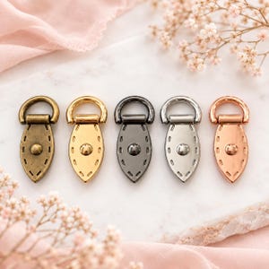 May include: Five metal bag connectors arranged in a row, each featuring a teardrop-shaped base with a central rivet and decorative stitching detail, topped with a D-ring. The finishes include antique brass, gold, gunmetal, silver, and rose gold.
