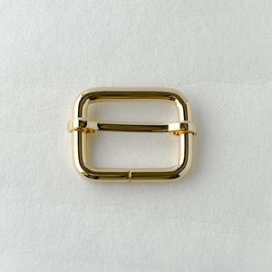 May include: Gold metal buckle with a rectangular shape. The buckle has a smooth, polished finish.