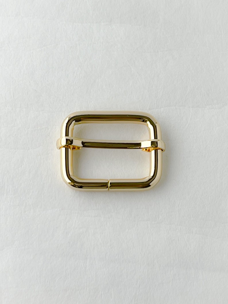 May include: Gold metal buckle with a rectangular shape. The buckle has a smooth, polished finish.