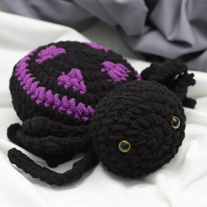 May include: A black crocheted spider with purple accents on its head and a smiling face. The spider has large black eyes with gold pupils.