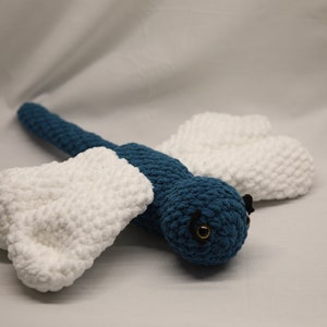 May include: A crocheted dragonfly toy with blue body and white wings. The dragonfly has black eyes and a gold button for a mouth.